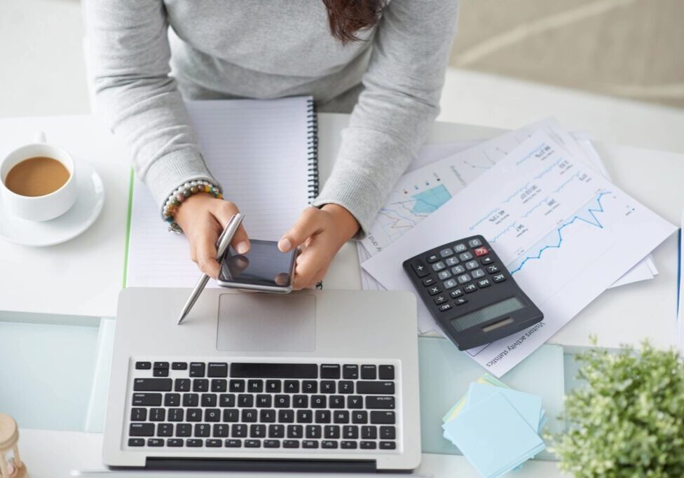 Person using smartphone with laptop, calculator, and financial documents nearby.